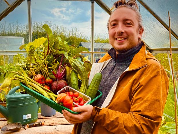 A member in a greenhouse holding a tray of beetroot, cucumbers and tomatoes he has just picked