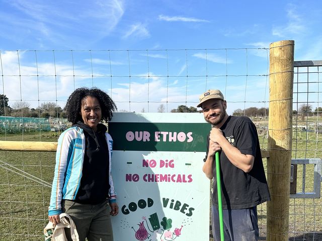Two members standing beside the painted site sign reading our ethos, no dig, no chemicals, good vibes